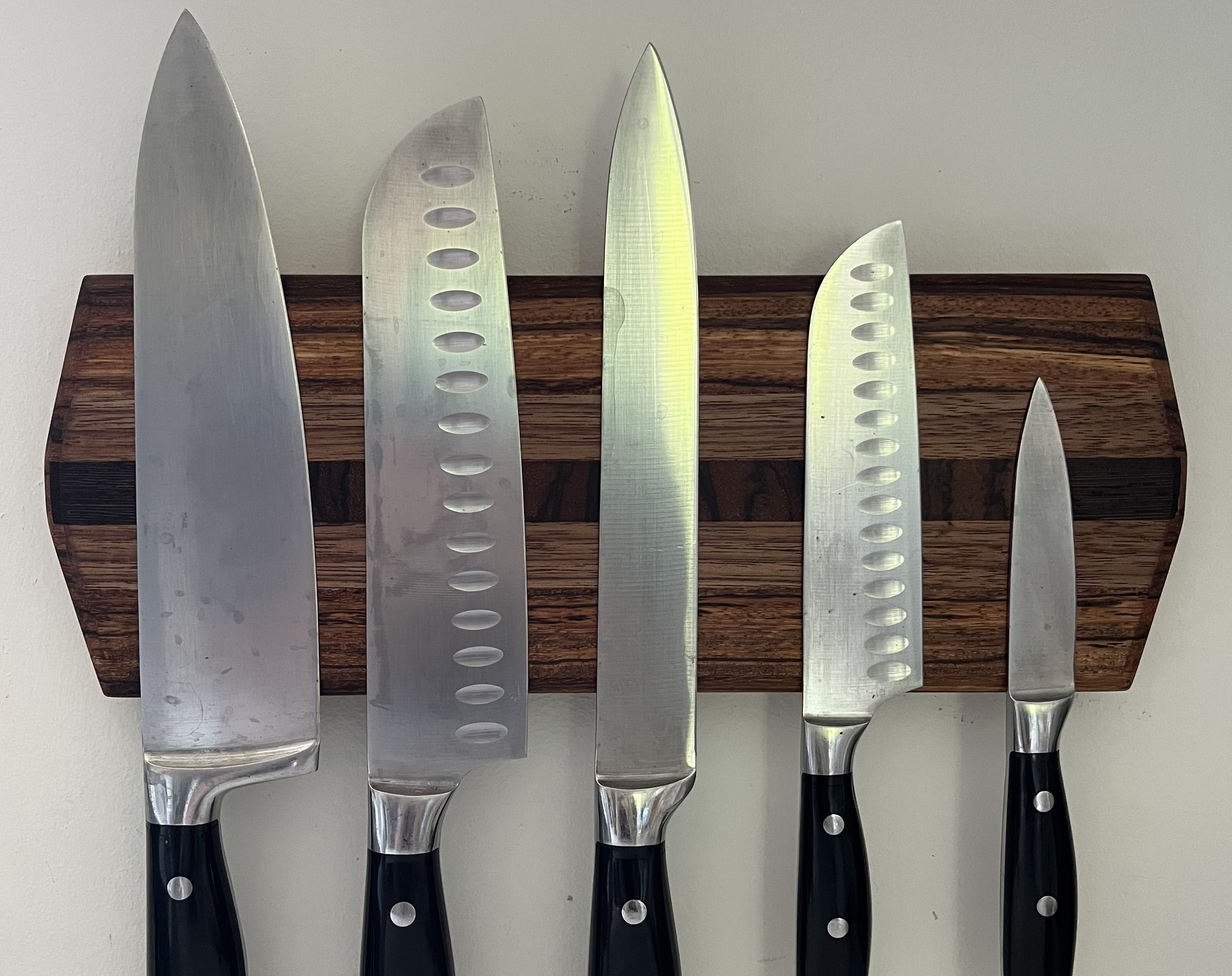 Wall-mounted magnetic knife holder made of walnut and wenge, mounted on a kitchen wall, holding multiple kitchen knives.