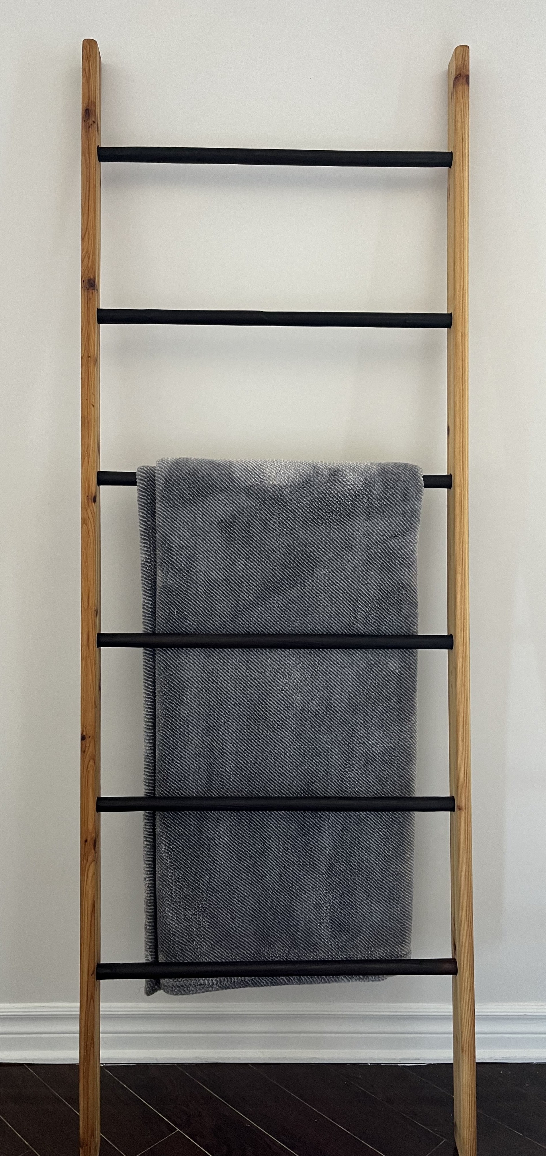 Leaning towel/blanket ladder with light spruce rails and black burnt dowel rungs, a grey towel hanging over the middle rungs